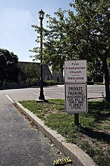 Photograph of a parking lot with two signs in the foreground: the sign above reads "First Presbyterian church: Welcome," the sign below says "PRIVATE PARKING: UNAUTHORIZED CARS WILL BE TOWED AWAY AT OWNER'S EXPENSE"