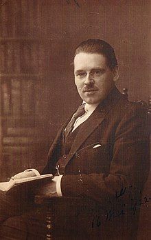 A man with rimless glasses, slicked back hair, and a toothbrush mustache in an ill-fitting suit sitting in a chair and holding a book