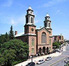 A brick church with two tall, symmetric steeples is seen in front of a city street, to the right of a wooded park.