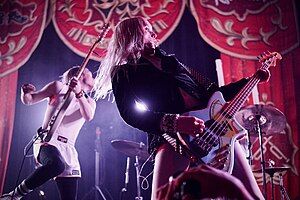 Angel (background) and Lulu (foreground) of Frost Children, playing the electric guitar during a climactic moment of a song. Illuminated by stage lights and backed by theatre curtains.