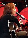 The side view of a woman with her eyes closed, singing into a microphone and playing a black guitar.