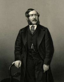 Three-quarter length portrait of white man with bushy side-whiskers and moustache, standing with head turned slightly to his left, his right hand resting on the back of a chair; well-dressed, wearing a dark open coat over buttoned waistcoat with watch chain, tie pin and ring shown