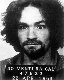 Black-and-white headshot photo of a crazy-eyed man with a dark mop of hair and beard