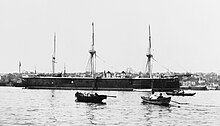 A large black warship sits in port with several small boats in the foreground.