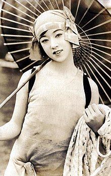 A young Japanese woman wearing a swimsuit and a cloche-style hat with large bows at the temples, holding a parasol