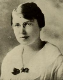 A young white woman with hair in a side-parted short style, wearing a white blouse with a wide collar