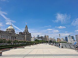 A wide curving walkway along a body of water to the right, crowded with many people and some large orange umbrellas underneath a sky colored by sunset. To the left of the walkway are light brownish-grey stone buildings in various late-19th and early-20th-century architectural styles, with taller buildings in the distance.