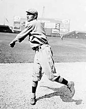 A man in a white baseball uniform and white baseball cap follows through after throwing a baseball on a grass field.