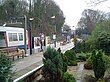 A grey railway platform with a series of light poles surrounded by brown trees, green bushes, and yellow and white flowers all under a white sky