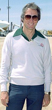 A grey-haired man wearing dark glasses, a cream sweater and jeans