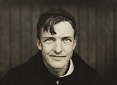 A dark-haired man wearing a black sweater and crownless baseball cap looks into the camera. His hair hangs down over his right side of his forehead, and he has a slight smirk on his face.