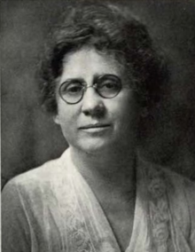 A middle-aged white woman wearing round glasses and a light-colored jacket and blouse; her dark hair is wavy