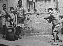 five children stand in-frame and another mostly off-frame; three are wearing no shirts, one of whom is swinging a stick at an airborne ball