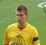 The head and upper body of young man wearing a yellow top, standing on a grass field. His top features a distinctive red, black and yellow crest, with the word "Watford" visible below it.