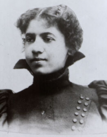 Carrie Still Shepperson, a Black woman with light skin, curly hair dressed with a center part, wearing a dramatic dark high-collared jacket with puffy sleeves and diagonal double-row button detail across chest