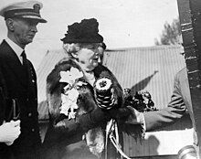 Black and white photograph of a men in a naval uniform jacket and white cap at the left, a women with a fur coat and hat at the centre