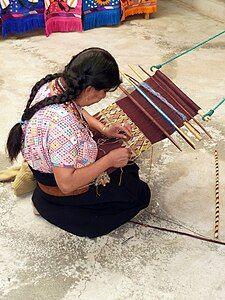 A loom made of sticks and string. The top endbar is tied to a fixed object using green rope; the lower end bar is attached to a leather strap around the weaver's back. Between, two heddle rods and several shedding rods. The sticks to one side are probably sword beaters. No shuttles or bobbins are being used.