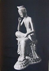 Three-quarter view of a seated statue in half-lotus position. The right foot rests on the left upper leg, the right elbow rests on the right knee with the right hand close to the head in contemplating pose. Black and white picture.