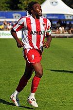 A young man wearing a red and white striped top, red shorts, red socks and white studded training shoes, standing on a grass field. The number 9 is visible on his shorts