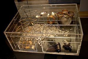 Transparent perspex box containing a shelf, two inner boxes, a large silver dish and many dozens of coins. The smaller inner box contains coins, whereas the larger contains two goblets and stacked ladles. The shelf holds gold bracelets, gold chains, and engraved spoons.