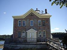 A brick building with yellow wooden trim and a pointed roof with a chimney and weather instruments on the roof. On the other side part of a lighthouse is visible. A wooden ramp leads up to a deck on its stone foundation surrounded by an iron fence.