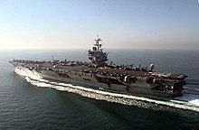 Aircraft carrier steaming away from camera in open sea. On deck is a large contingent of aircraft