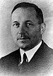 Head of a middle-aged white man with thinning hair, wearing a dark suit coat, white shirt, and dark tie.