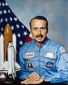 man with mustache in astronaut uniform, model of space shuttle to side, U.S. flag in background