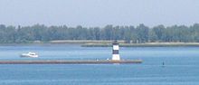 A pier with a square, black and white-striped lighthouse at the end. A small motorboat is paralleling the pier, heading away from the lighthouse.