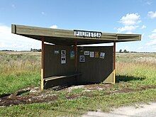 A wooden bus stop with two walls making a triangular shape, which is painted green, has a triangular roof, and has a bench.