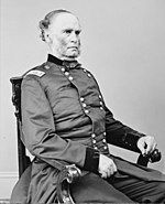 Black and white photo shows a man with a receding hairline and mutton chops. He is seated and wearing a dark uniform with two rows of buttons.