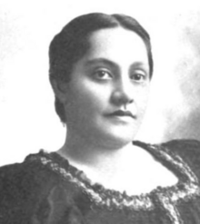 A young Hawaiian woman, dark hair parted center and dressed to the nape, wearing a dark dress with a square ruffled collar