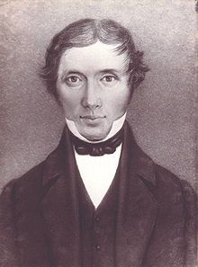 A young clean-shaven man with wavy hair, wearing dark coloured jacket and vest over a white shirt with a high collar. He is looking straight ahead, with a faint smile