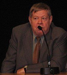 Color photo: Medium shot of Arto Paasilinna, sitting behind a table, speaking into a microphone