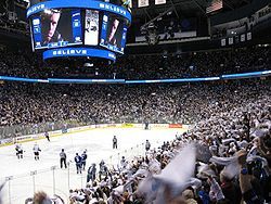 An arena set up for ice hockey. Players skate towards center ice, while a larger crowd is waiving white towels. Over head is a jumbo-tron with an extreme close-up of a player looking seriously at the camera, above and below the picture is the word "Believe" in white lights with a blue light background.