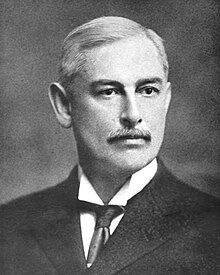 A black-and-white photographic portrait of an older, mustached man from the shoulders up, wearing a suit, with a necktie