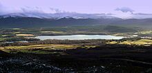 A relatively small loch on the River Spey is surrounded by forest and moorland. In the background can be seen the Cairngorm Mountains, the summits of which are covered with snow.