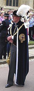 An elderly gentleman walking along. He is wearing a suit with grey trousers, over which is draped a heavy robe and a gold change. A black hat with a large white plume sits on his head. In the background stands a police woman separated from a group of people by a white cordon.