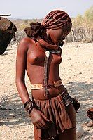 As is customary in Himba culture and climate, a Himba girl of northern Namibia wears a traditional skirt made from calfskin leather, headdress and jewelry which signify her social status.