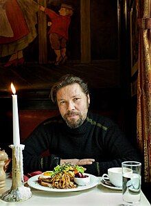 middle-aged white male wearing dark top, sitting at a dining table, facing camera with head slightly tilted