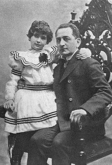 A white man with his daughter, about ten years old, seated on his lap; she is wearing a white dress with a wide collar and black striped embellisments; he is wearing a suit