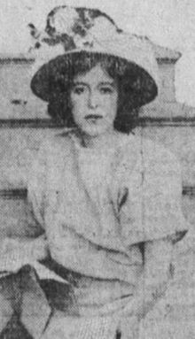 A young white woman with dark hair, wearing a light-colored dress and a brimmed hat