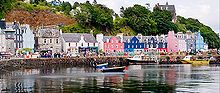 A row of brightly coloured stone buildings front a harbour with a tree-clad cliff behind. The houses are painted predominantly in shades of pink, blue and white. In the foreground, small boats lie in the water by the shelter of a stone pier.