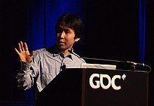 A Japanese artist man gestures in front of a podium.