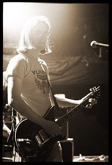 black-and-white, washed out image of Steven Wilson standing onstage in front of a microphone in a light-coloured print t-shirt, playing an electric guitar