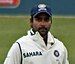 A man in the white Indian test cricket jersey, wearing a blue hat. In the background is the field of a cricket ground.