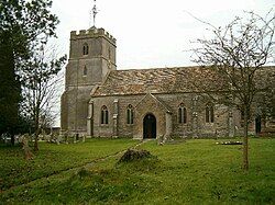 Brown stone building with square tower at the left hand end. In the foreground is a grass area with gravestones.