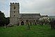 A square base of five steps made from large stones, supporting a base and a modern cross in front of a low stone church and tower