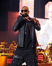 A black man wearing sunglasses and a dark outfit performs with a microphone