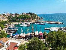 An harbor with multiple trees and buildings on the land, and numerous boats docked behind a breakwater, two small ships on the open sea are in the background